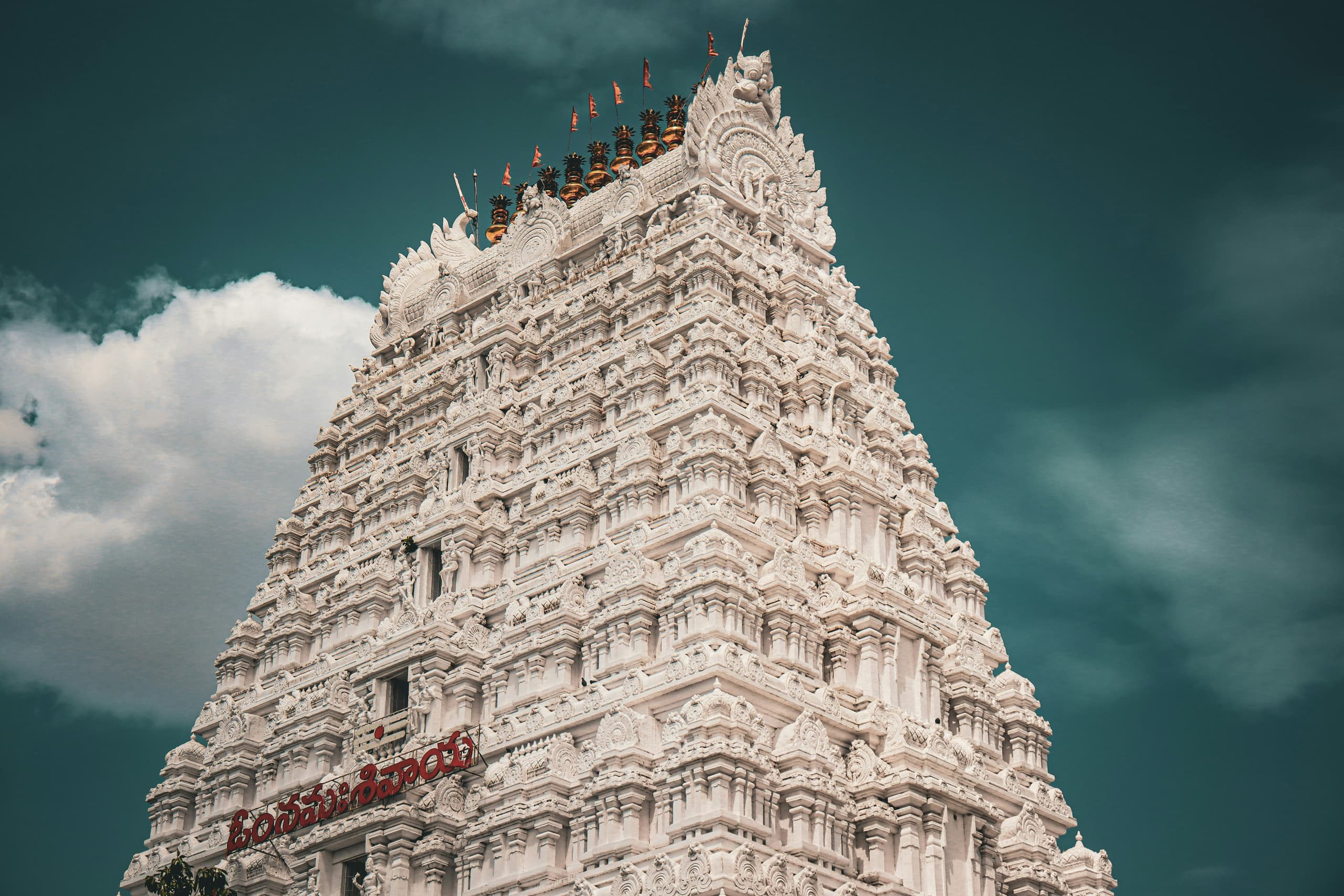 A white temple gopuram crowned with golden kalashas beneath a stormy teal sky